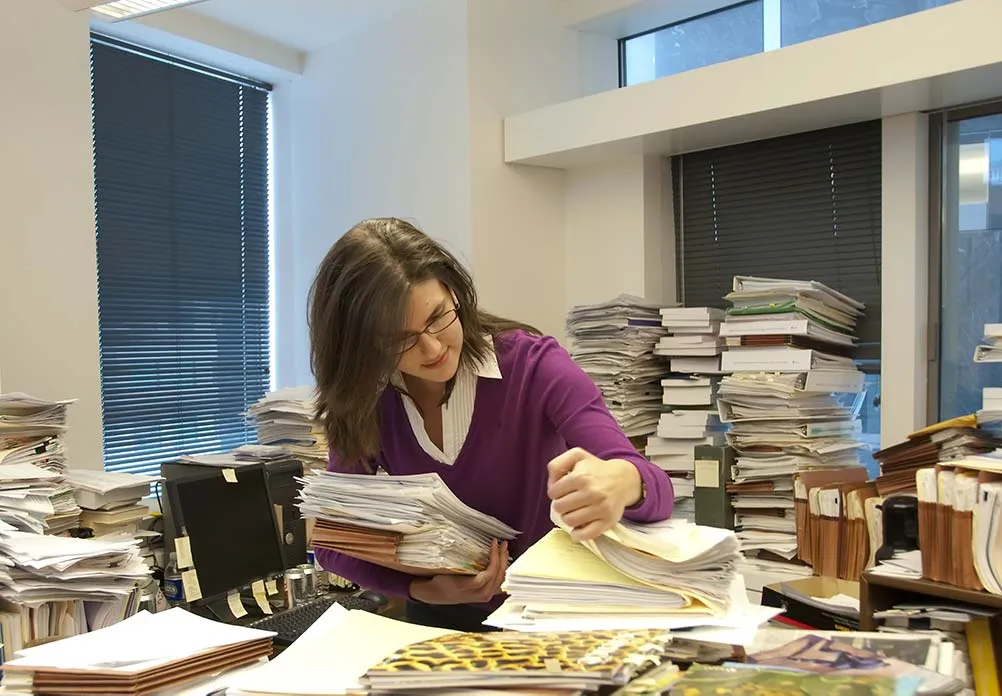 Photo de référence : une femme trie des piles de dossiers sur un bureau encombré, devant des archives empilées
